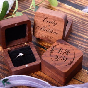 May include: Three wooden ring boxes with engraved text. The first box says "Emily & Matthew 08.07.25". The second box says "Eternal Love E M 08.14.2023". The third box is open and has a velvet interior with a ring inside.