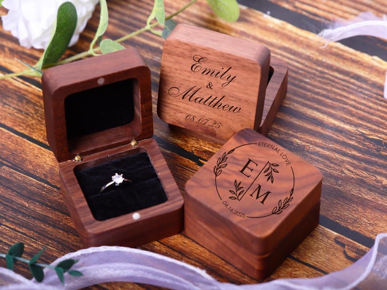 May include: Three wooden ring boxes with engraved text. The first box says "Emily & Matthew 08.07.25". The second box says "Eternal Love E M 08.14.2023". The third box is open and has a velvet interior with a ring inside.