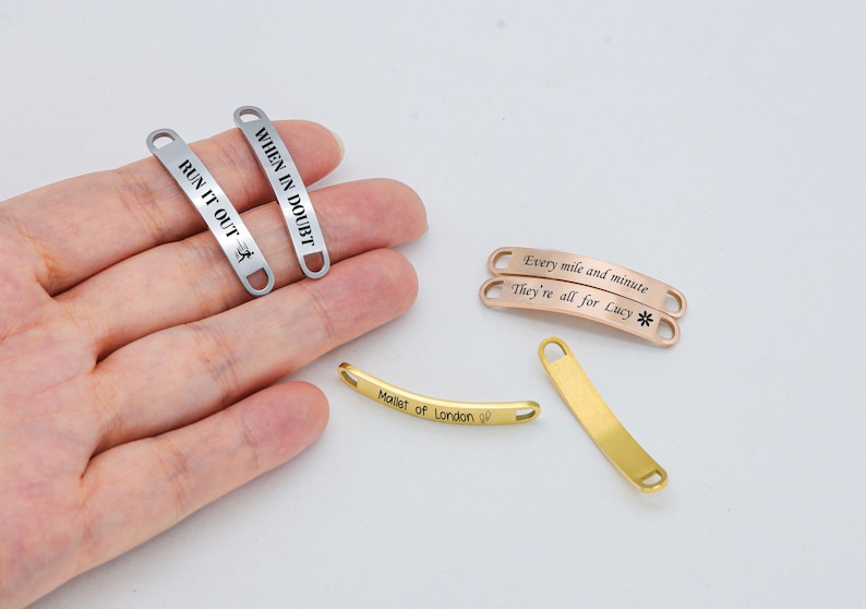 May include: Five silver and gold metal tags with engraved text. The tags are curved and have rounded ends. The text on the tags reads: "WHEN IN DOUBT", "RUN IT OUT", "Every mile and minute", "They're all for Lucy", and "Mallet of London".