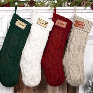 Puede incluir: Cuatro calcetines navide&ntilde;os de punto trenzado en verde, blanco, rojo y beige, cada uno con una etiqueta de nombre de cuero personalizada. Los calcetines cuelgan de una repisa blanca decorada con vegetaci&oacute;n y adornos festivos.