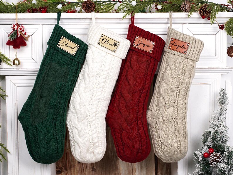 Puede incluir: Cuatro calcetines navide&ntilde;os de punto trenzado en verde, blanco, rojo y beige, cada uno con una etiqueta de nombre de cuero personalizada. Los calcetines cuelgan de una repisa blanca decorada con vegetaci&oacute;n y adornos festivos.