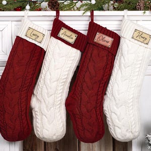Puede incluir: Cuatro calcetines navide&ntilde;os de punto trenzado cuelgan de una repisa blanca. Dos son rojos, dos son blancos, y cada uno tiene una etiqueta de nombre de madera personalizada. Los nombres son Larry, Amelia, Oliver y Henry. Decoraci&oacute;n festiva.
