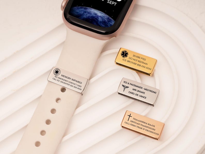 May include: Medical alert tags for a smartwatch, featuring engraved text and symbols. One tag is attached to a cream watch strap. Other tags are gold, silver, and rose gold. The tags provide vital medical information.