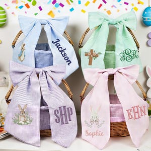 May include: Four Easter baskets with seersucker bows and personalised details. Two baskets have purple and white striped bows, one with "SHP" and the other with a bunny and "Sophie". The other two have blue and green bows with names and embroidered designs.