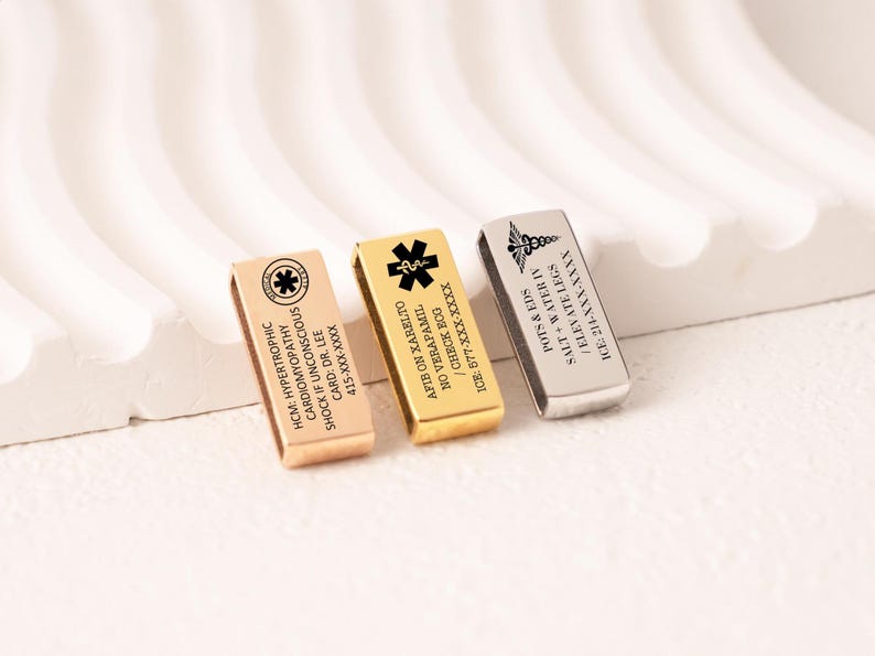 May include: Three rectangular medical alert bracelets in rose gold, gold, and silver. Each bracelet has engraved text with medical information and symbols. The bracelets are displayed on a white, textured surface.