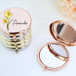 Könnte beinhalten: Ein Kompakt-Spiegel mit roségoldener Oberfläche und einem floralen Design. Der Spiegel hat einen weißen Hintergrund mit einer gelben Narzissenblüte und dem Namen "Amanda" in schwarzer Schrift. Der Spiegel ist geschlossen und das Design ist auf der Vorderseite sichtbar.