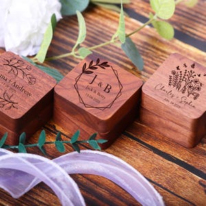 May include: Three wooden ring boxes with engraved floral designs and personalized names and dates. The boxes are made of dark wood and have a square shape.
