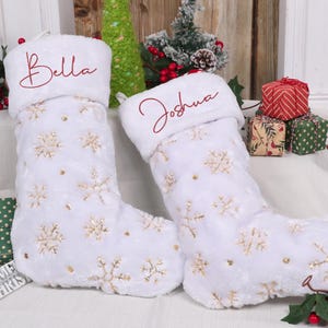 Peut inclure: Deux bas de Noël blancs avec des détails de flocons de neige et de paillettes. Le haut de chaque bas est brodé d'un nom en script rouge. Des cadeaux et de la verdure sont en arrière-plan.