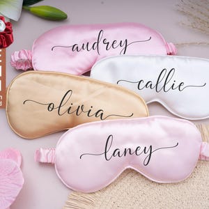 May include: Several satin sleep masks in shades of pink, cream, and gold, each bearing a name in cursive script. The image also shows matching scrunchies and text that reads "FREE BUY 1 GET 1".