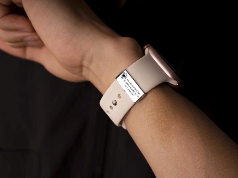 May include: A light pink smartwatch with a matching strap is worn on a wrist. A silver rectangular tag is attached to the strap, displaying text and a circular graphic. The watch is set against a dark background.