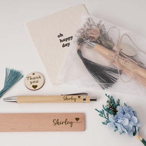 May include: A selection of personalised gifts, including a wooden pen, bookmark, and tag, all engraved with the name "Shirley." A pen is wrapped in a clear bag with dried flowers. A small bouquet of blue flowers is also included.