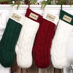 Puede incluir: Cuatro calcetines navide&ntilde;os de punto trenzado en verde, blanco y rojo cuelgan de una repisa blanca. Cada calcet&iacute;n tiene una etiqueta de madera con el nombre: "Larry", "Amelia", "Oliver" y "Henry". Decoraci&oacute;n festiva.