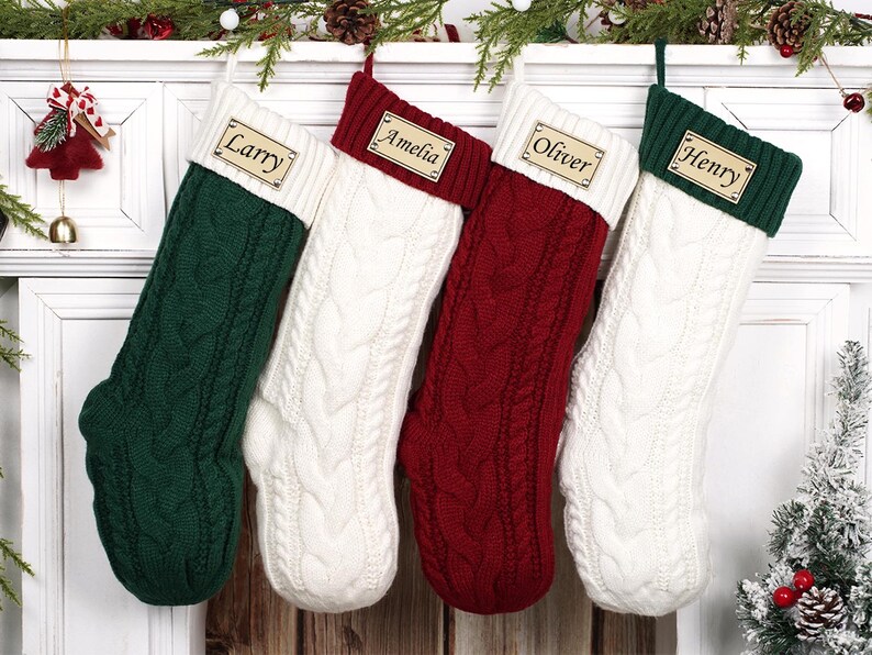 Puede incluir: Cuatro calcetines navide&ntilde;os de punto trenzado en verde, blanco y rojo cuelgan de una repisa blanca. Cada calcet&iacute;n tiene una etiqueta de madera con el nombre: "Larry", "Amelia", "Oliver" y "Henry". Decoraci&oacute;n festiva.