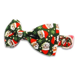 May include: A festive Christmas-themed dog bow tie. The bow tie features a dark green background with a pattern of Santa Claus faces, red hats, and white snowflakes. The collar is red with a silver buckle.