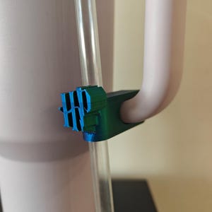 Puede incluir: Primer plano de un portavasos de plástico azul y verde fijado a un vaso de color morado claro. El portavasos tiene un diseño texturizado y sostiene una pajita de plástico transparente.