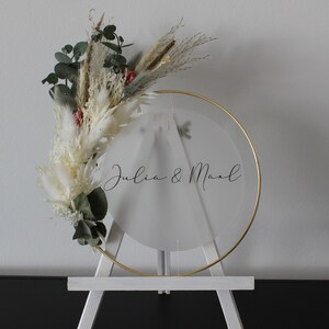 Könnte beinhalten: Ein weißer Staffelei mit einem goldenen Metallring, der einen durchsichtigen Acrylkreis mit dem Text "Julia & Mark" in schwarzer Kalligraphie hält. Der Ring ist mit getrocknetem weißem Pampasgras und Eukalyptus verziert.
