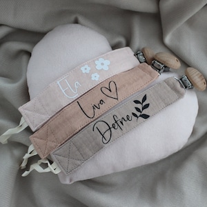 Könnte beinhalten: Drei rosa Stoff-Schnullerketten mit Holzclips. Die Schnullerketten sind mit den Namen "Ela", "Liva" und "Defne" in schwarzer Schrift personalisiert. Die Schnullerkette mit dem Namen "Ela" hat außerdem eine weiße Blumenstickerei.