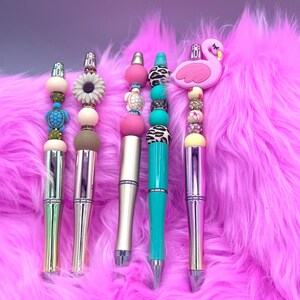 Może przedstawiać: Cztery kolorowe długopisy z koralikami i różnymi ozdobami tematycznymi. Długopisy leżą na różowym, puszystym podłożu. Ozdoby obejmują żółwia, słonecznik, różową i białą muszlę oraz różowego flaminga.