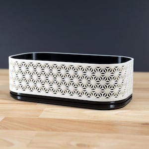 Op de afbeelding: Een rechthoekige opbergcontainer met een geometrisch sterrenpatroon. De container is wit met een zwart interieur en basis. De container is ongeveer 20 cm lang, 10 cm breed en 8 cm hoog.