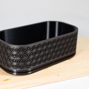 Op de afbeelding: Een zwarte rechthoekige container met een geometrisch sterrenpatroon aan de zijkanten. De container is gemaakt van een donker materiaal en staat op een lichtgekleurd houten oppervlak. De binnenkant is leeg.
