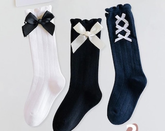 Peut inclure: Trois paires de chaussettes hautes avec des nœuds de différents designs. Une paire est blanche avec un nœud noir, une paire est noire avec un nœud blanc et une paire est bleu marine avec un nœud blanc en forme de croix.