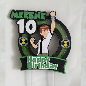 May include: A birthday cake topper with a cartoon character design. The topper features the name "Mekene" and the number "10" in green and white. The words "Happy Birthday" are displayed on a black banner, with a green and black circular design.