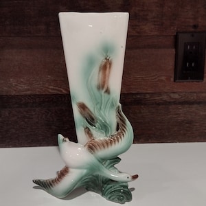 Puede incluir: Un jarrón de cerámica blanca con un diseño verde y marrón de juncos estilizados y dos pájaros. El jarrón es alto y delgado con una abertura rectangular.