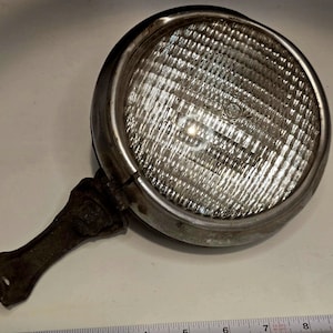 May include: An antique, round headlight with a chrome bezel and a textured glass lens. The headlight has a metal bracket and handle. A measuring tape is placed below the headlight.