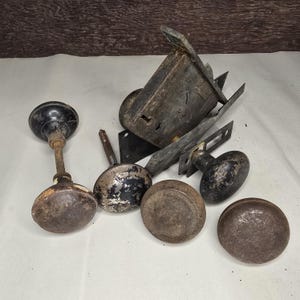 May include: A collection of five weathered, dark-toned metal door knobs with varying degrees of paint wear and oxidation, accompanied by a large, rectangular, heavy-duty iron mortise lock mechanism with a visible keyhole and internal latch components.