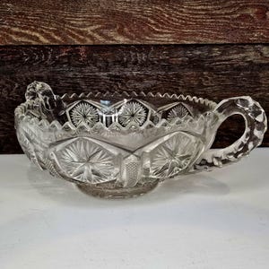 May include: Clear, cut-glass serving bowl with a handle. The bowl features a starburst and geometric pattern, with a scalloped rim. The handle is also cut with a textured design. The bowl is likely vintage and is sitting on a white surface.