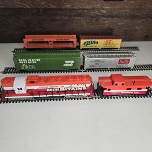 May include: A collection of toy train cars and engines on tracks. The set includes a red and white engine with "COMIN' ROUND THE MOUNTAIN" text, and various freight cars in orange, green, yellow, and silver. The cars have different company names.