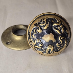 May include: A round metal door knob featuring a black finish with worn gold-toned highlights and intricate raised scrollwork patterns, accompanied by a separate circular brass mounting plate with a central opening and screw holes.
