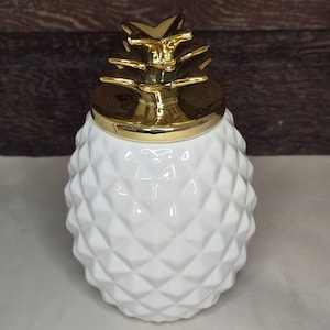 May include: A white ceramic jar shaped like a pineapple with a textured diamond pattern on the body. The lid is metallic gold and features a sculpted pineapple leaf design at the top, serving as a handle for the removable lid of the container.