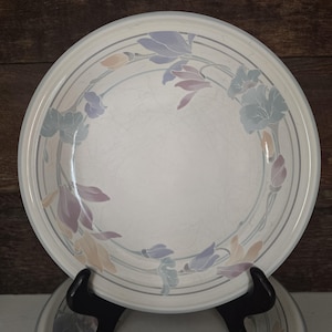 May include: A white ceramic dinner plate with a floral design. The plate features a light gray border with two concentric gray lines. The floral pattern includes lavender, peach, and light blue flowers. The plate is displayed on a black stand.
