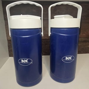 May include: Two blue insulated water jugs with white lids and handles. Each jug has a white logo with the letters "AK" inside an oval. The jugs are cylindrical and appear to be made of plastic.