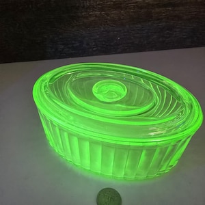 Könnte beinhalten: Eine leuchtend grüne, ovale Glasschale mit Deckel. Die Schale hat ein geripptes Design an den Seiten und einen kreisförmigen Knopf auf dem Deckel. Das Glas leuchtet in einem lebendigen Neonfarbton, was auf Vintage oder Uranglas hindeuten könnte.