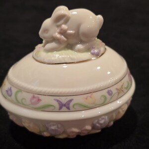 May include: A cream-colored ceramic Easter egg-shaped trinket box with a bunny figurine on top. The box is decorated with pastel flowers, butterflies, and a gold trim. The bunny has a pink bow.