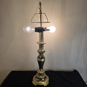 May include: A vintage brass table lamp with a decorative base and a two-bulb design. The lamp features a detailed, ornate design and a black shade holder. The bulbs are lit, casting a warm glow.