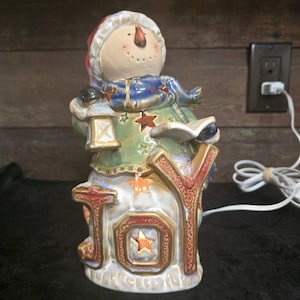 May include: A ceramic snowman figurine holding a lantern and a book, standing on a base that spells out "JOY" in colorful letters. The snowman is wearing a red hat, a blue scarf, and a green coat. The figurine is lit up with warm white lights.
