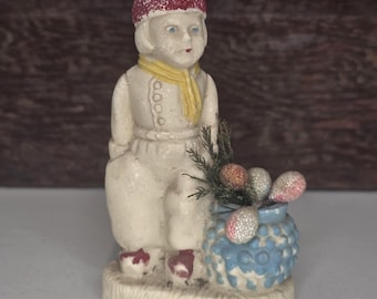 Puede incluir: Figura vintage de una persona sentada con gorro rojo, bufanda amarilla y atuendo blanco. La figura sostiene un jarrón azul con elementos florales decorativos. La figura tiene una base color crema y probablemente sea de cerámica.