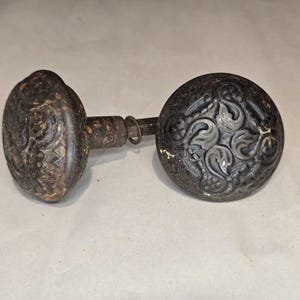 May include: A pair of dark, weathered metal door knobs featuring a raised, intricate scrollwork pattern on the circular faces. The knobs are connected by a central square metal spindle with a small metal ring attached to the shaft.