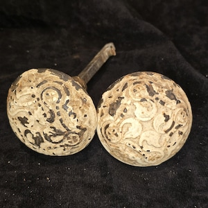 May include: Two round metal door knobs featuring a distressed white finish with exposed dark metal underneath. The knobs have a raised, intricate scrollwork pattern across the surface. One knob includes a square metal spindle extending from the back.