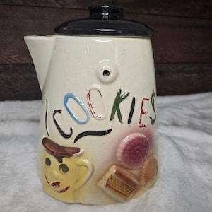 May include: A vintage ceramic cookie jar shaped like a jug with a black lid. The jar is decorated with colourful cartoon cookies and the word "COOKIES" in a whimsical font.