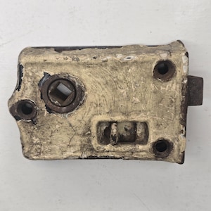 May include: An antique door latch with a distressed, off-white finish. The metal latch has a rectangular shape with a square opening for the spindle and several screw holes. The latch shows signs of age and wear.