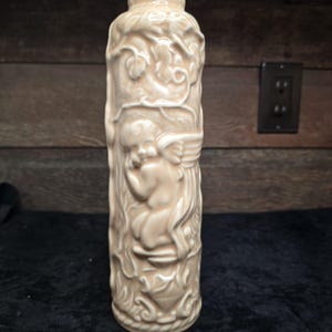 May include: A ceramic vase with a cream-colored glaze features a sculpted cherub with wings. The vase is decorated with a vine pattern.