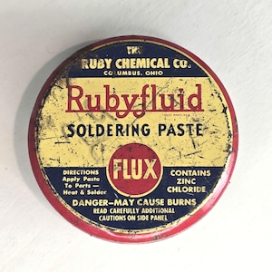 May include: A round, vintage Rubyfluid soldering paste tin. The tin has a red rim, a yellow center, and blue text. The text includes "Rubyfluid", "Soldering Paste", and "Flux". It also warns "Danger-May Cause Burns".