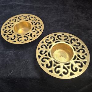 May include: Two brass candle holders with intricate, openwork designs. Each circular holder features a central cup for a candle and a decorative pattern of swirling cutouts around the edge. The candle holders are displayed on a dark surface.