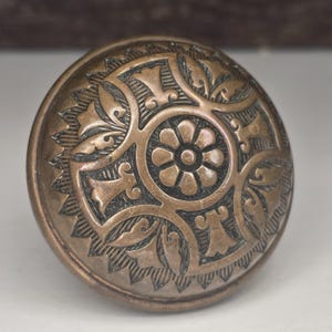 Könnte beinhalten: Ein bronzefarbener, runder Schrankknopf mit einem verzierten Blumen- und Blattdesign. Der Knopf hat eine zentrale Blume, die von detaillierten Mustern umgeben ist. Die Kanten haben ein strukturiertes, gewelltes Design.