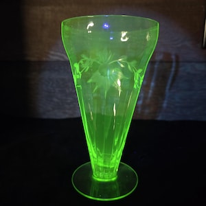Op de afbeelding: Een groene glazen vaas met een gegraveerd bloemenmotief aan de zijkant. De vaas heeft een uitlopende bovenkant en een brede basis.