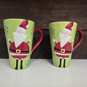 Può includere: Due tazze in ceramica verde con manici rossi. Ogni tazza presenta un design di Babbo Natale 3D con barba bianca e vestito rosso. Le tazze hanno uno sfondo verde con disegni di fiocchi di neve bianchi.
